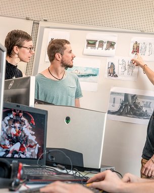 Vier Personen stehen um eine Wand mit Konzeptzeichnungen. Ein Mann zeigt auf eine Zeichnung, während die anderen aufmerksam zuschauen. Im Vordergrund sind Computer und Kopfhörer sichtbar.