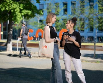 Two women are smiling on the street as a person rides by on an electric scooter. One woman is holding a smartphone.