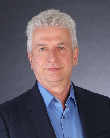 Ein Mann mit grauen Haaren trägt einen dunklen Anzug und ein hellblaues Hemd. Er blickt direkt in die Kamera vor einem neutralen Hintergrund.