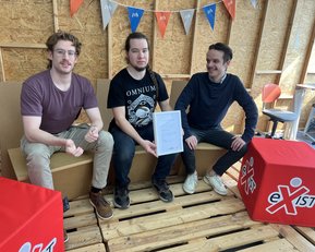 Three men are sitting on a wooden bench in a room adorned with colorful flags. The man in the middle is holding a document while the other two are looking at him. Red dice with the word 'eXist' are visible in the foreground.