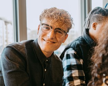 A young man with curly blonde hair and glasses is sitting at a table. He is wearing a dark blue shirt and smiling. In the background, there are other people and windows visible.