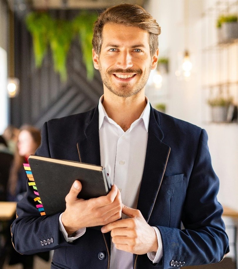 Ein Mann in einem blauen Blazer hält ein Notizbuch und lächelt. Im Hintergrund sind unscharfe Personen in einem Café zu sehen. Die Atmosphäre wirkt freundlich und einladend.