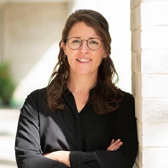 A woman with glasses stands against a wall, smiling with her arms crossed in front of her chest. She is wearing a black shirt.