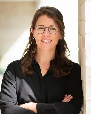 Eine Frau mit Brille steht lächelnd an einer Wand, die Arme vor der Brust verschränkt, in einem hellen, modernen Raum.