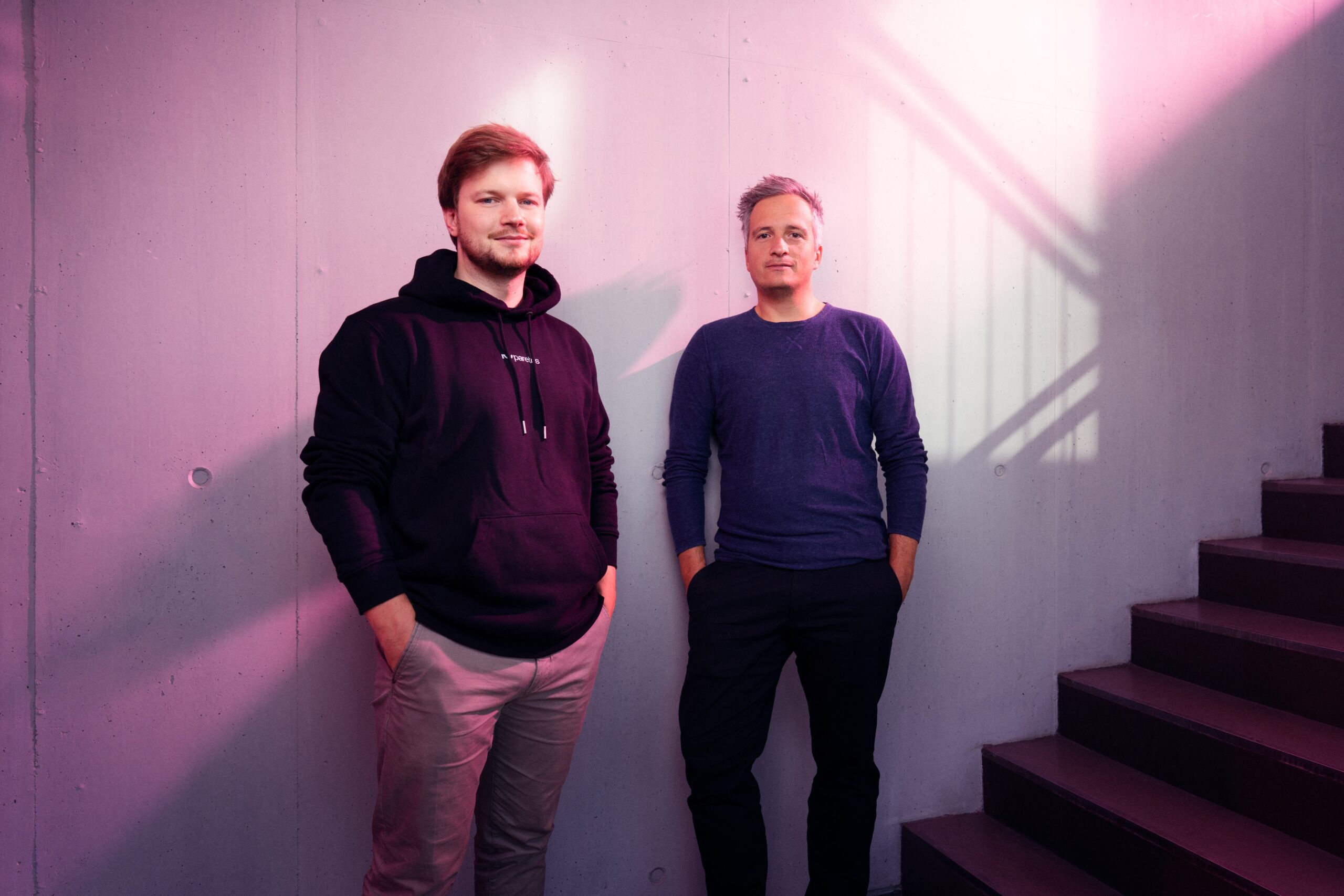Zwei Männer stehen nebeneinander vor einer Wand mit sanften Lichtreflexen. Der Mann links trägt einen schwarzen Hoodie, der Mann rechts ein blaues Oberteil. Im Hintergrund sind Treppenstufen sichtbar.
