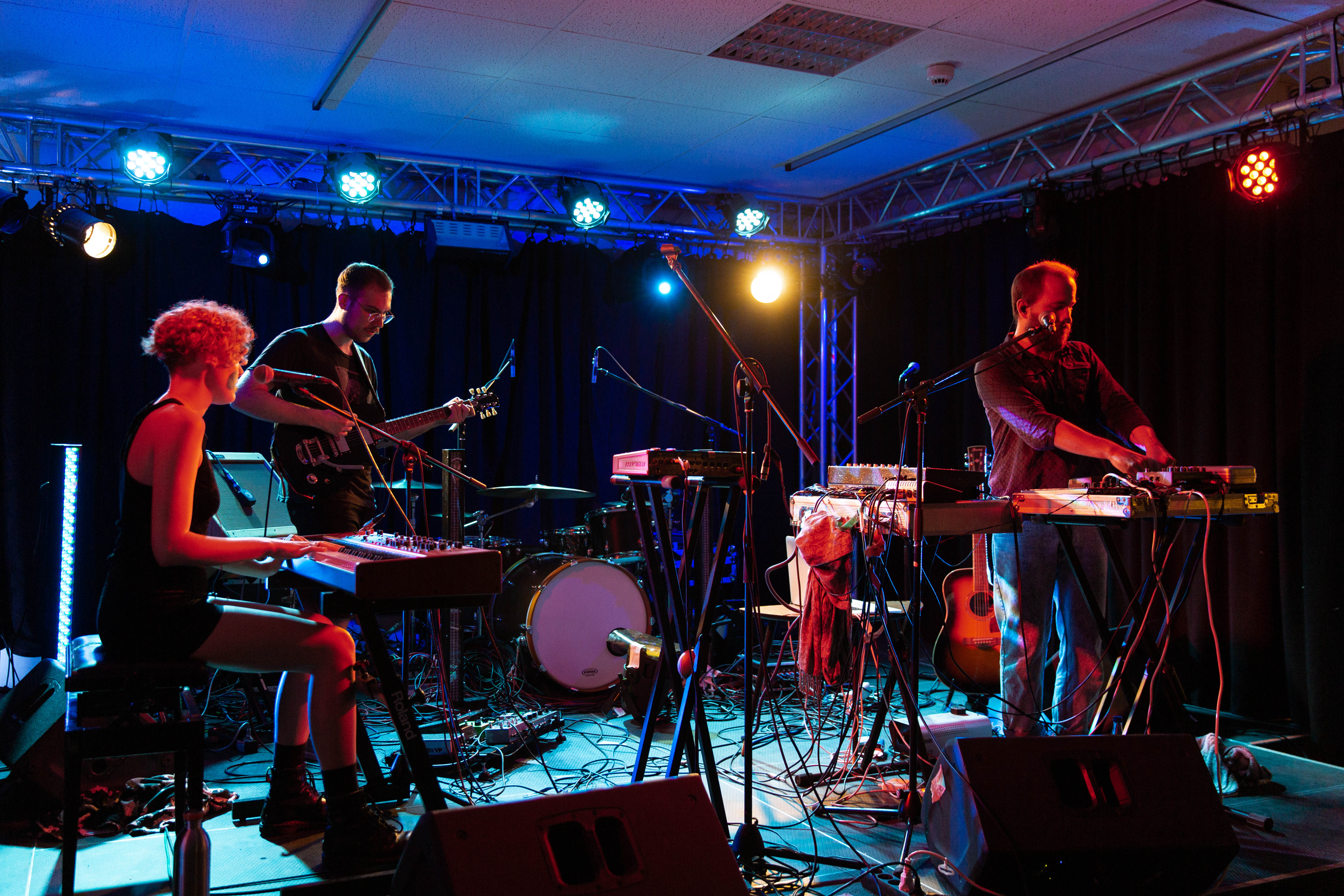 Drei Musiker auf einer Bühne, einer spielt Gitarre, einer an einem Synthesizer, und eine Frau am Keyboard. Bunte Lichter beleuchten die Szene.
