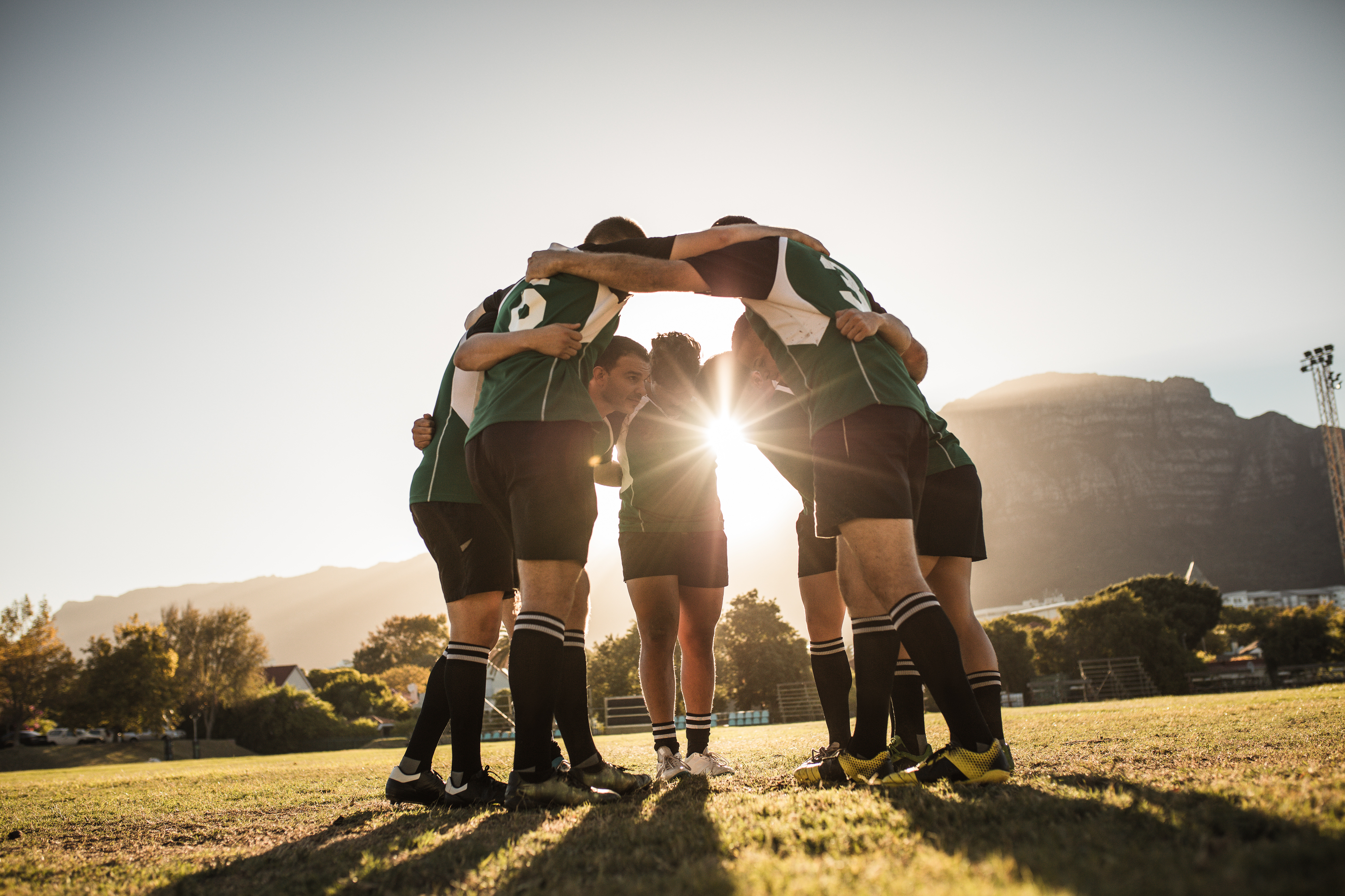 Eine Gruppe von Rugbyspielern steht in einem Kreis und umarmt sich, während die Sonne hinter ihnen aufgeht.