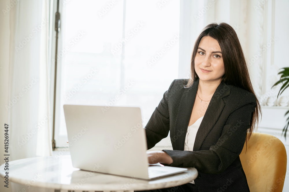 Eine Frau in einem schwarzen Blazer sitzt an einem Tisch mit einem Laptop. Sie schaut direkt in die Kamera und wirkt selbstbewusst. Im Hintergrund sind Fenster und Pflanzen zu sehen.
