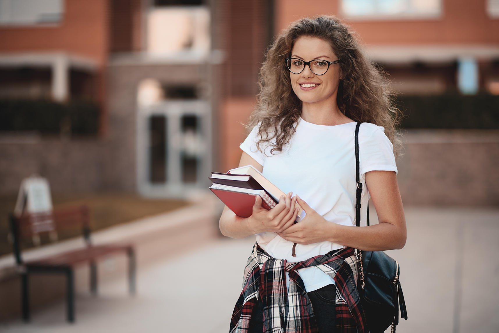 Eine Frau mit lockigem Haar und Brille hält mehrere Bücher in der Hand und trägt ein kariertes Hemd um die Taille.