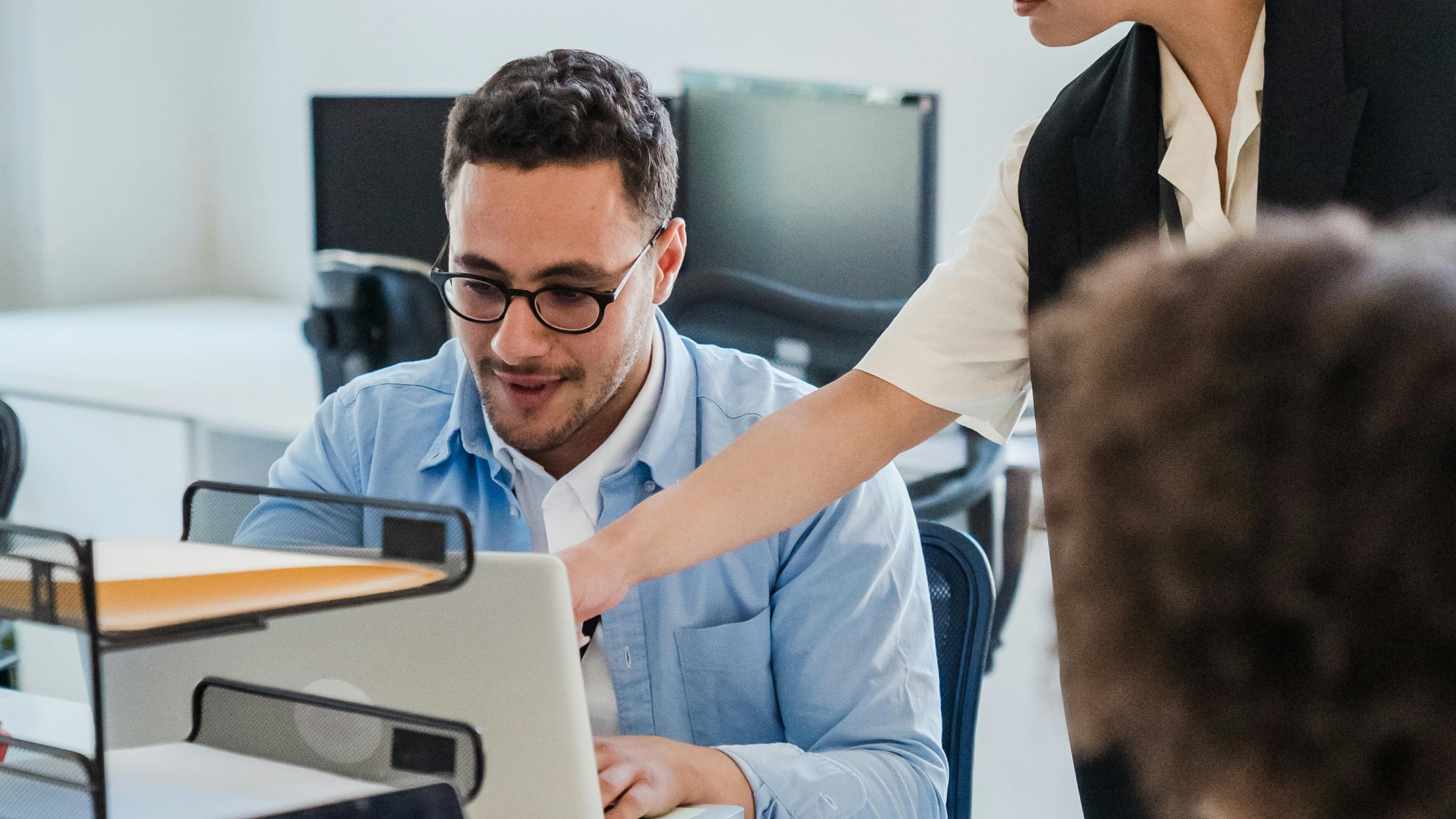 Ein Mann sitzt an einem Computer, während eine Frau ihm etwas zeigt. Beide sind in einem modernen Büro mit mehreren Arbeitsplätzen.