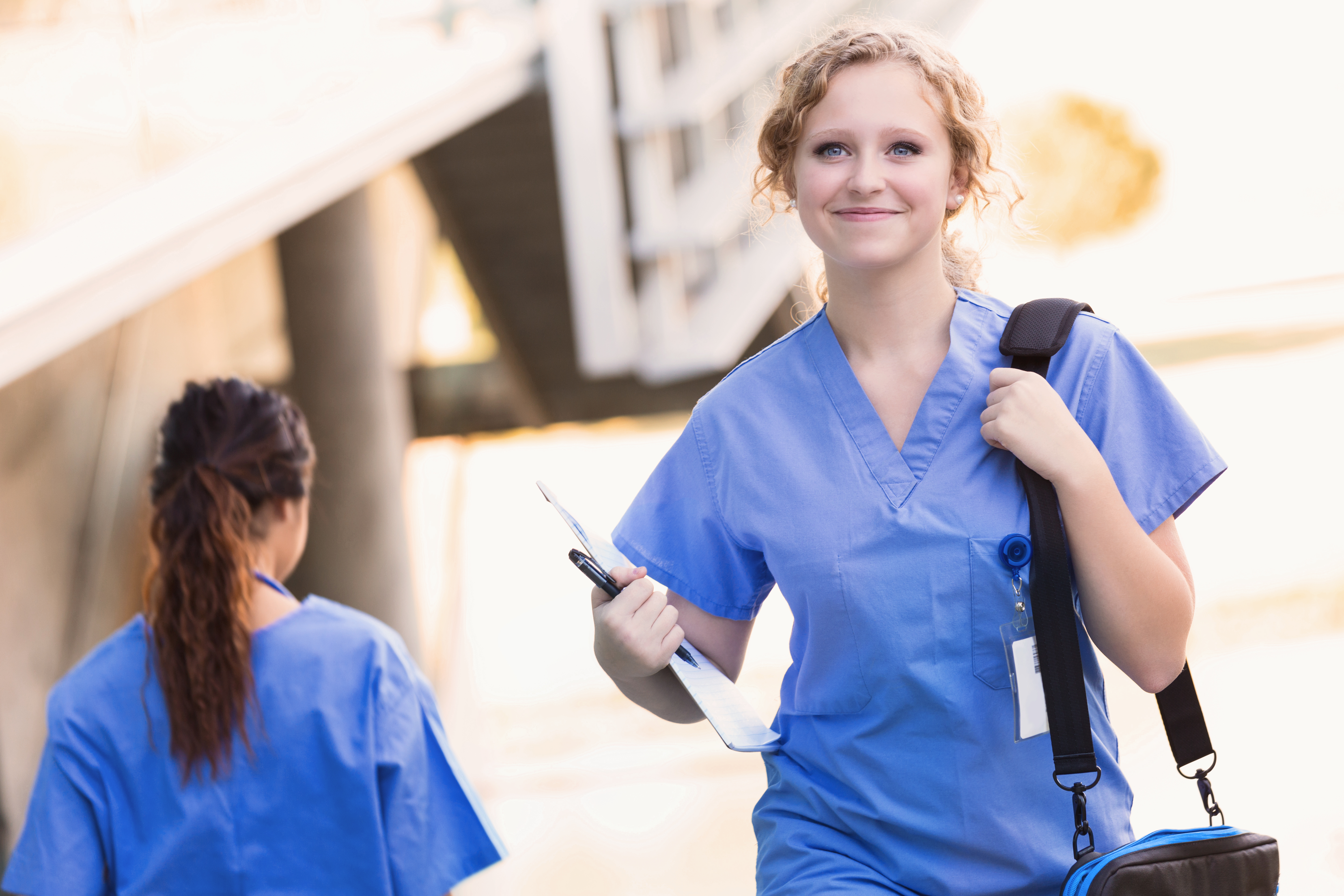 Eine lächelnde Krankenschwester in blauer Arbeitskleidung trägt eine Tasche und ein Clipboard, während sie auf einem Weg geht. Eine weitere Person in ähnlicher Kleidung ist im Hintergrund zu sehen.