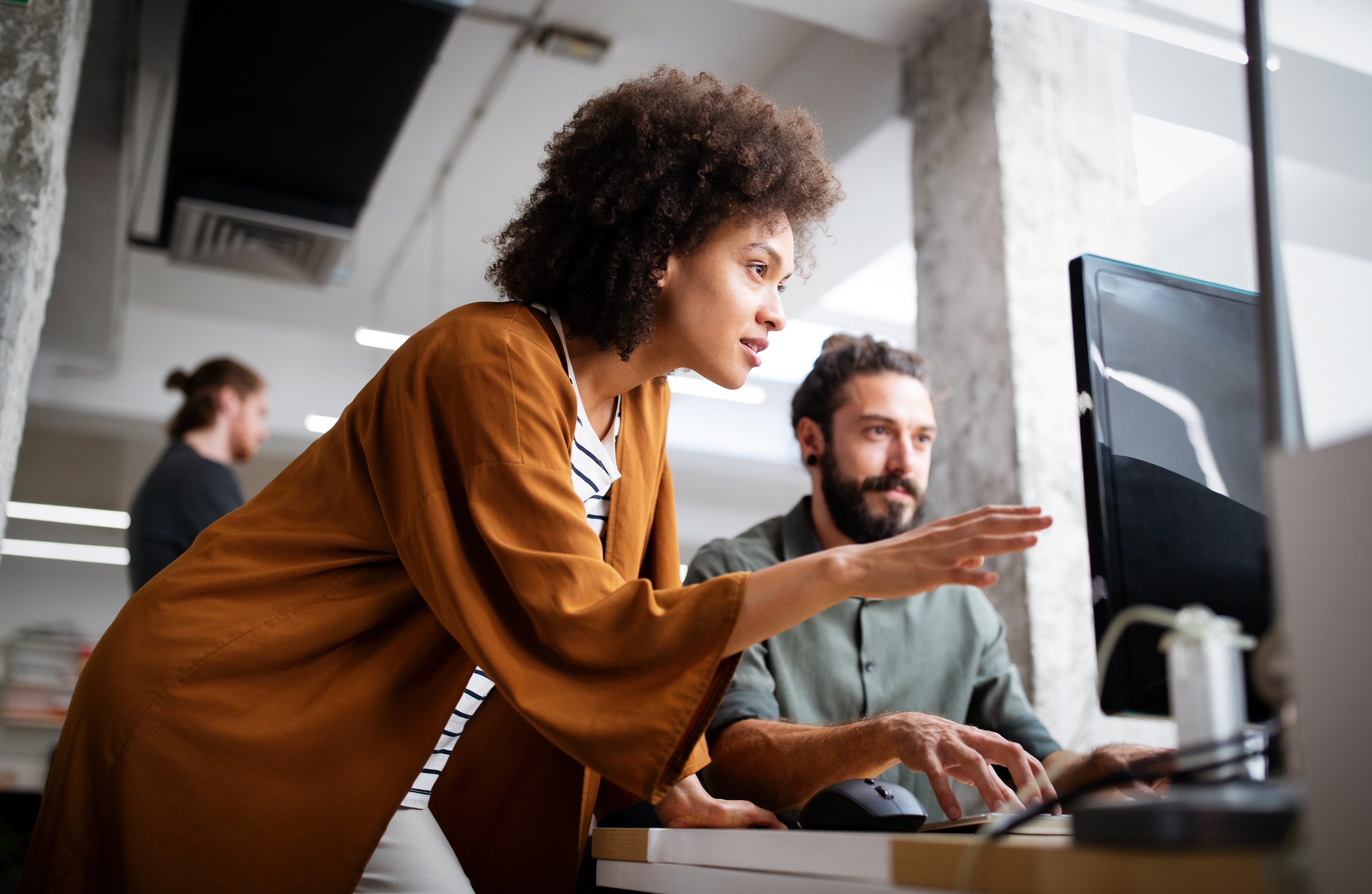 Eine Frau mit lockigem Haar erklärt etwas einem Mann am Computer in einem modernen Büro. Im Hintergrund sind weitere Personen zu sehen.