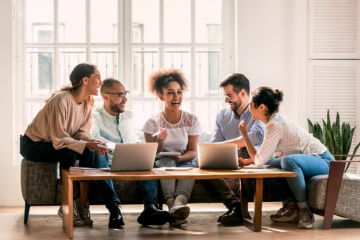 Sechs Personen sitzen um einen Tisch mit Laptops, lachen und diskutieren in einem hellen Raum mit großen Fenstern.