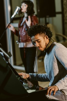 Ein Musiker spielt Klavier, während eine Sängerin im Hintergrund singt. Beide sind in einem modernen Studio mit Mikrofon und entspannter Atmosphäre.