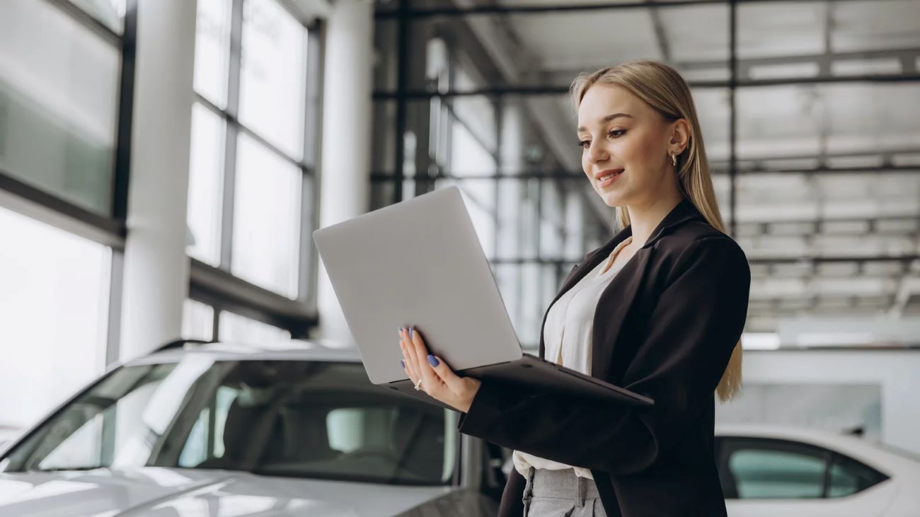 Eine junge Frau in einem Anzug steht in einem modernen Autohaus und arbeitet an einem Laptop, während sie lächelt. Im Hintergrund sind Autos und große Fenster zu sehen.