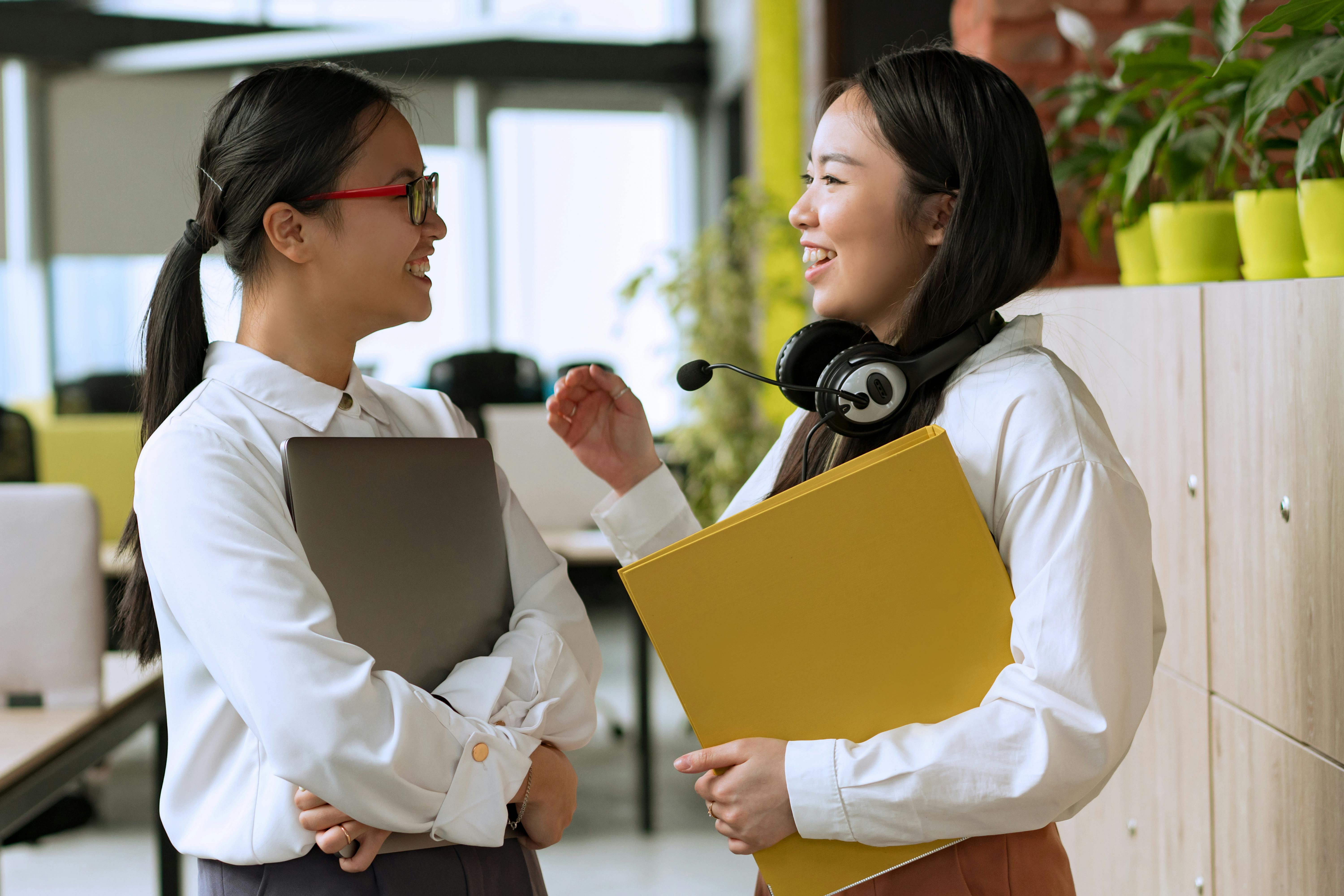 Zwei Frauen im Büro sprechen miteinander, eine hält einen Laptop und die andere eine gelbe Mappe. Beide tragen formelle Kleidung.