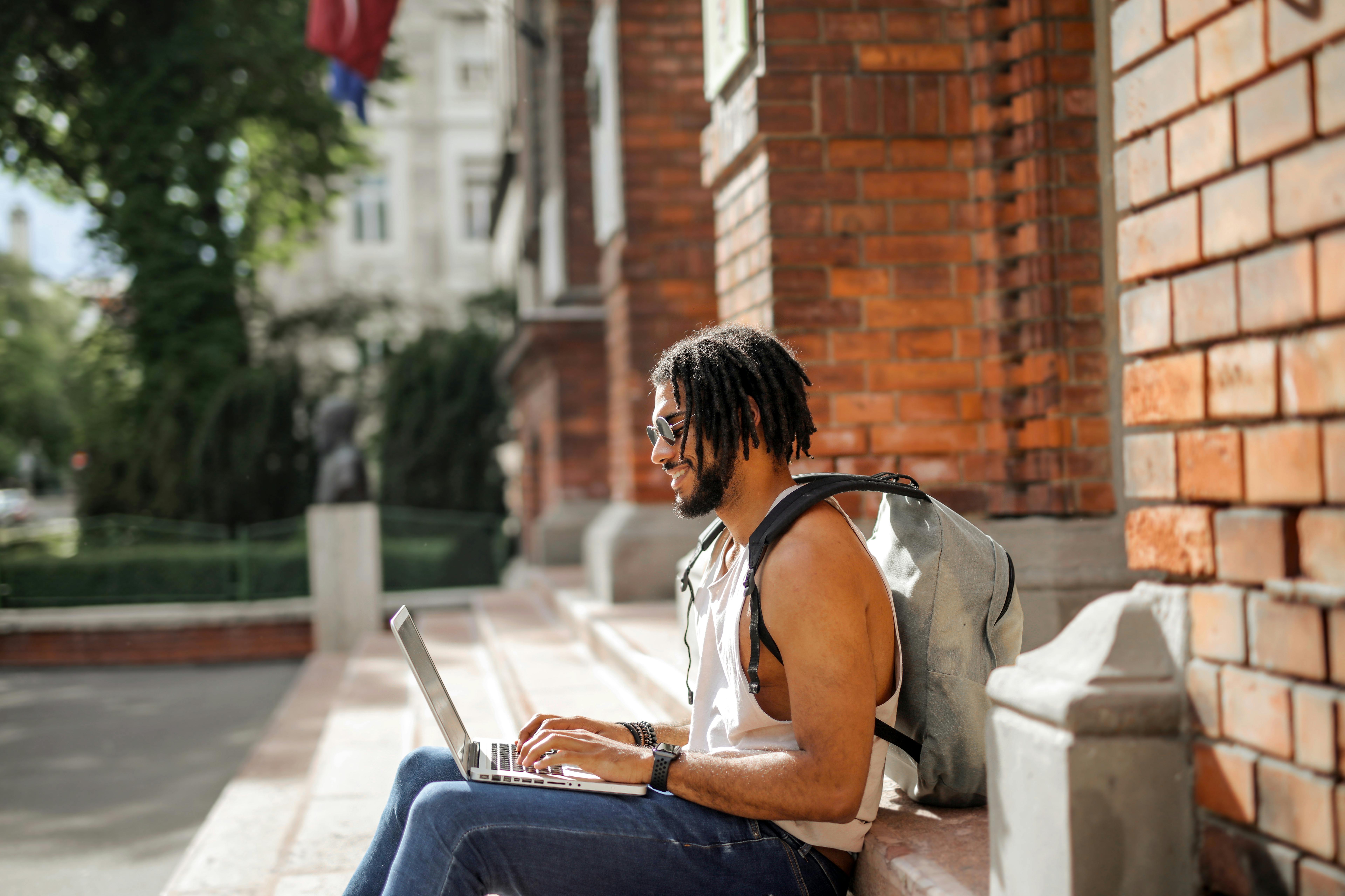 Ein Mann mit Dreadlocks sitzt auf einer Treppe und arbeitet an einem Laptop, während er einen Rucksack trägt.