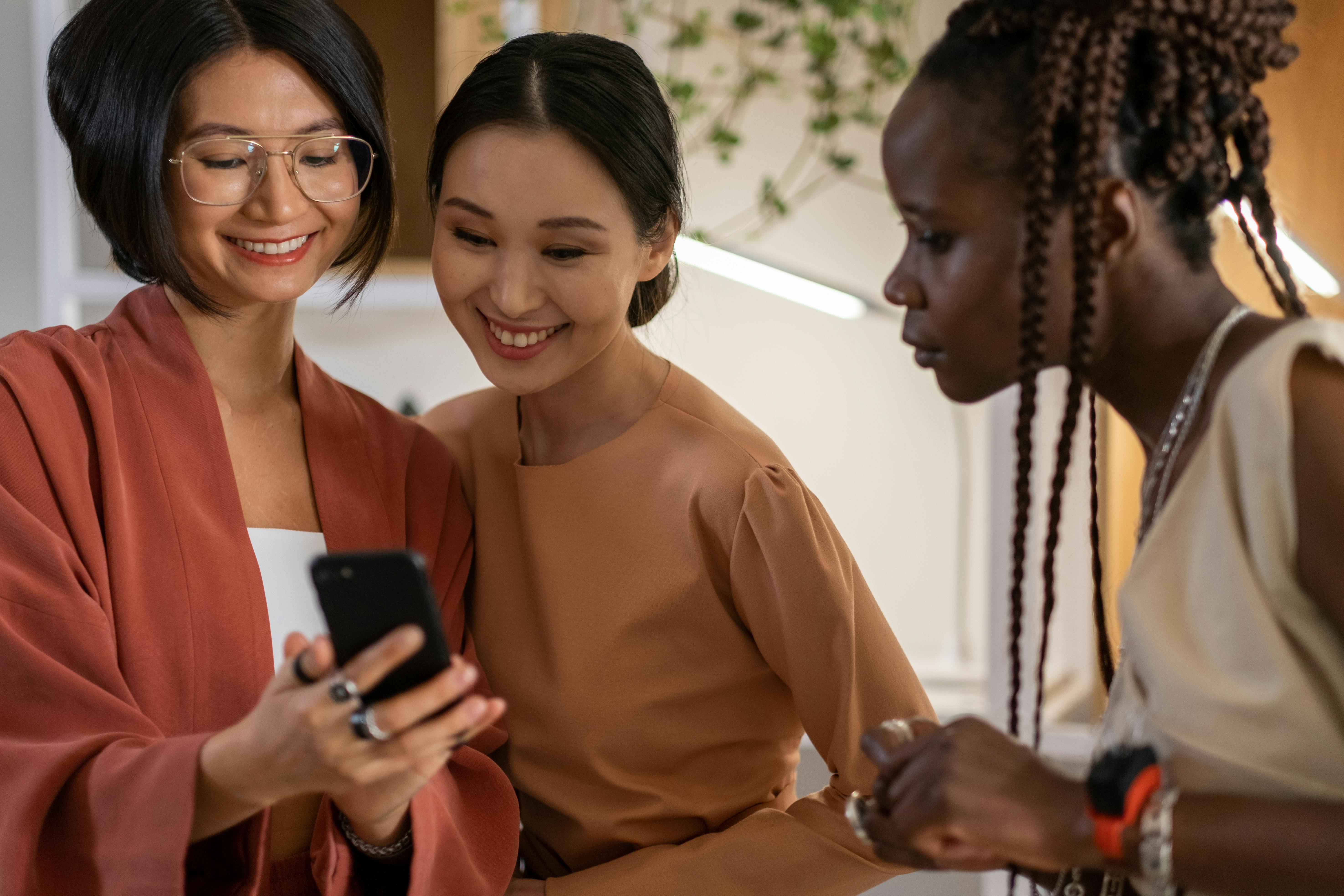 Drei Frauen betrachten gemeinsam ein Smartphone, während sie lächeln. Eine Frau trägt eine orangefarbene Jacke, die anderen sind in neutralen Tönen gekleidet.