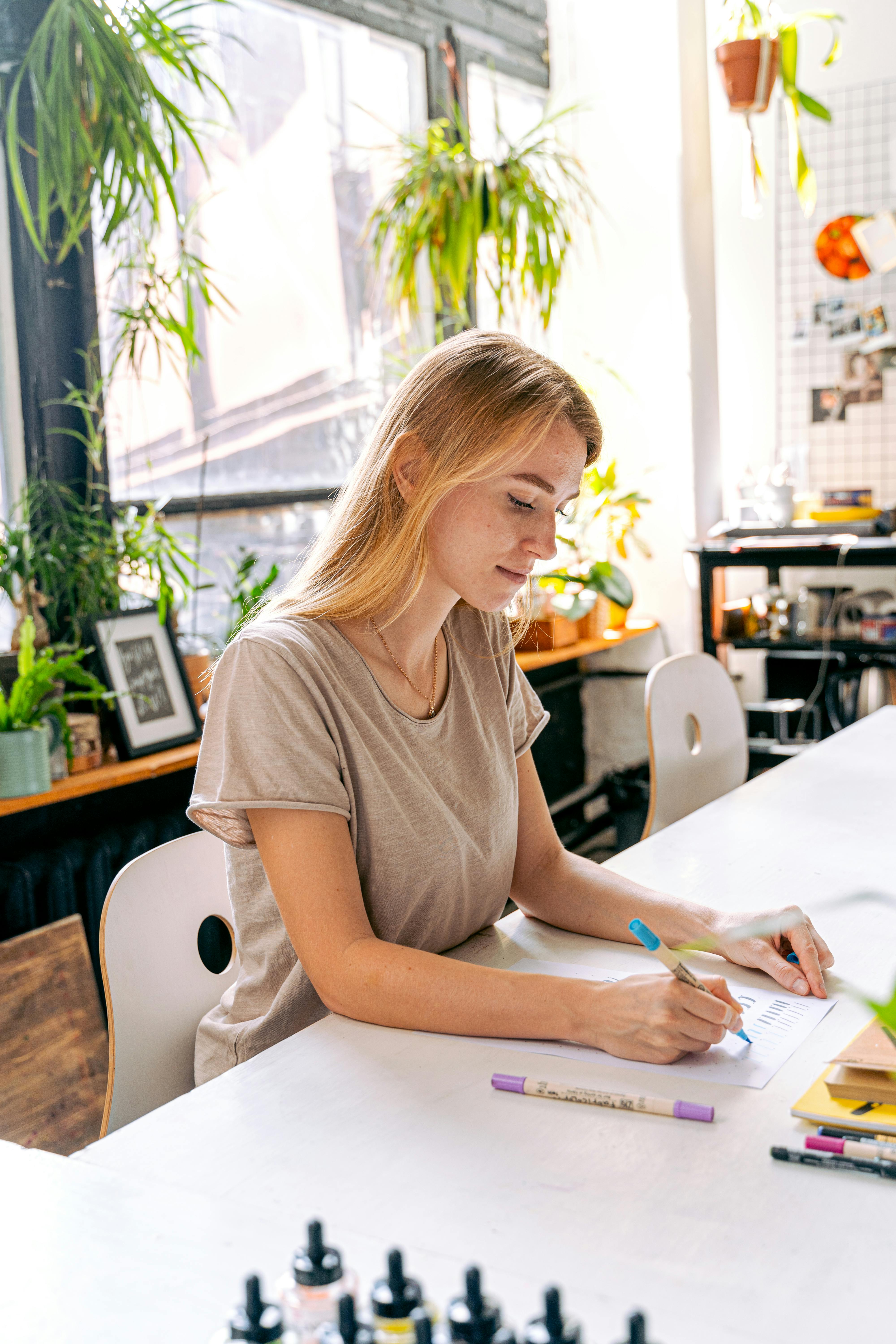 Eine junge Frau sitzt an einem Tisch und zeichnet mit einem Stift auf Papier, umgeben von Pflanzen und kreativen Materialien.