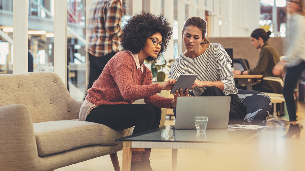 Zwei Frauen sitzen auf einem Sofa und betrachten gemeinsam ein Tablet, während ein Laptop auf dem Tisch steht.