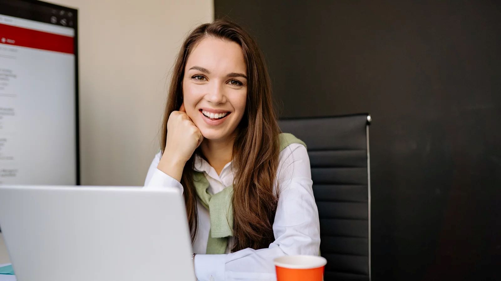 Eine lächelnde Frau sitzt an einem Schreibtisch mit einem Laptop und einer Tasse Kaffee. Im Hintergrund ist ein Bildschirm mit Text zu sehen. Sie trägt ein weißes Hemd und einen grünen Pullover.